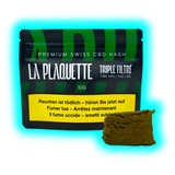 Im Vordergrund steht eine rechteckige Verpackung mit der Aufschrift 'LA PLAQUETTE TRIPLE FILTRÉ', die als Premium Swiss CBD Hasch beworben wird. Die Verpackung ist überwiegend grün mit schwarzen und weißen Schriftzügen. Darunter befindet sich ein kleiner brauner Block, der das Produkt, CBD Hasch, zeigt. Die Verpackung enthält weitere Hinweise in mehreren Sprachen, darunter einen Warnhinweis zum Rauchen. Ein leuchtend blauer Rahmen hebt die Verpackung und das Produkt hervor.